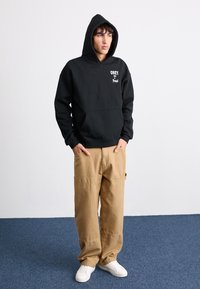 Black hooded sweatshirt with white "OBEY" text, kangaroo pocket, paired with tan baggy pants and white sneakers on textured blue floor.