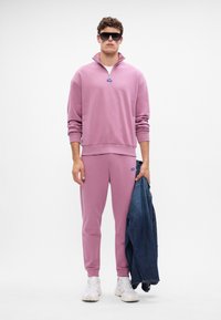 Svetlo roza flis pulover s polovičnim zipom in rebrastimi manšetami, v kombinaciji s usklajenimi trenirkami. Model drži denim jakno, nosi sončna očala in superge.