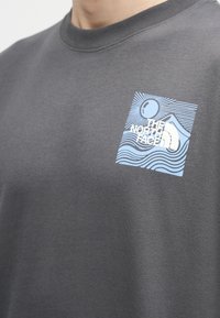 Gros plan sur un t-shirt gris avec un logo The North Face bleu et blanc représentant un soleil, des vagues et des montagnes sur la poitrine.