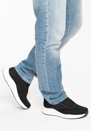 Person trägt hellblaue Jeans und schwarze Sportschuhe mit weißen Sohlen, steht mit einem leicht angehobenen Fuß.