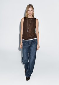 Bruine mouwloze top in laagjes over een witte shirt, wijde blauwe jeans en bruine schoenen, met een lange zilveren hangerketting.