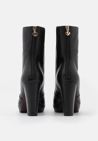 Botas de cuero negro hasta el tobillo con un tacón cuadrado alto, con una cremallera dorada con un charm en forma de corazón en la parte de atrás y una textura lisa.