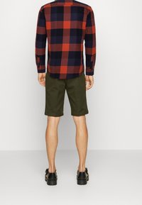 Hombre vestido con una camisa de manga larga de cuadros en negro y rojo, pantalones cortos de tiro a la rodilla en color verde oliva y sandalias gruesas negras, de pie contra un fondo blanco.