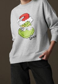 Sweat-shirt gris avec un graphique coloré du Grinch portant des lunettes de soleil, un chapeau de Père Noël rouge et une bordure en fourrure blanche. Coupe décontractée avec poignets côtelés.