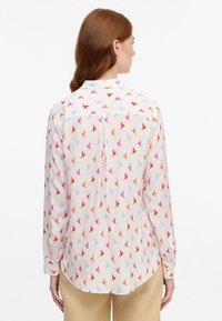 Long-sleeve shirt in cream fabric with a multicolored bird pattern in red, pink, and blue. Features a collar and buttoned cuffs.