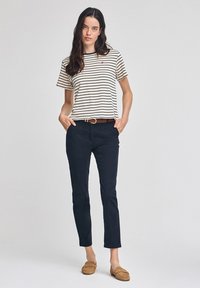 Randig beige och marinblå t-shirt, marinblå byxor, brun bälte, tan moccasiner; modellen står självsäkert med händerna i fickorna mot en neutral bakgrund.