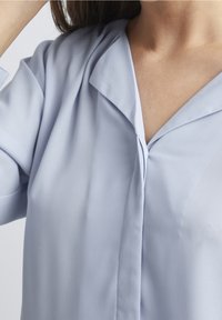 Blusa celeste chiaro con una consistenza morbida, dotata di colletto pieghettato, dettagli a plissé verticali e un design frontale senza bottoni.