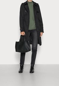 Schwarzer Trenchcoat über einem grünen Pullover, kombiniert mit schwarzen Jeans und Ankle Boots. Hält eine strukturierte schwarze Handtasche mit zwei Griffen.