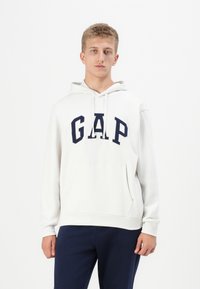 Sweat à capuche blanc en tissu doux avec un logo "GAP" bleu marine, poche kangourou et poignets côtelés, offrant une coupe décontractée.