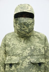 Kamuflāžas raksts parka zaļos toņos, aprīkota ar kapuci, sejas masku un divām priekšējām kabatām ar spiedpogu aizdari.