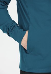 Person som bär en teal långärmad skjorta med högra handen delvis i en sidficka mot en neutral bakgrund.