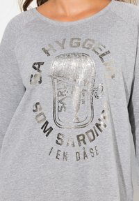 Top gris de manga larga con un gráfico de lata de sardinas metálica y texto, cuello redondo y mangas raglán. Tejido suave y texturizado.