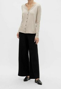 Beige långärmad cardigan med v-ringning, knäppning och dekorativa knappar på ärmarna, i kombination med vida svarta byxor och svarta loafers.