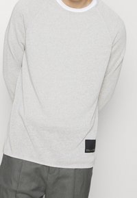 Pull gris clair texturé avec un col rond, doté de manches raglan et d'une petite étiquette noire sur le côté inférieur gauche.