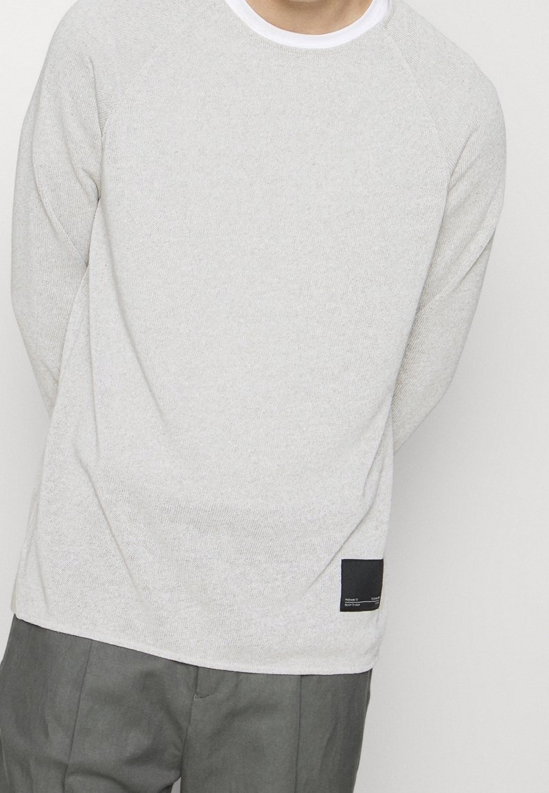 Pull gris clair texturé avec un col rond, doté de manches raglan et d'une petite étiquette noire sur le côté inférieur gauche.