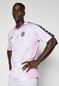 Zawodnik ubrany w biało-różową koszulkę piłkarską z czarnymi markowymi paskami na ramionach, stojący z rękami na biodrach na jednolitym tle.