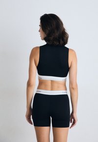 Crna sportska brusa s visokim izrezom i širokim trakama, u kombinaciji s crnim kratkim hlačama. Oboje imaju bijelu traku s "Calvin Klein" logotipom.