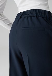 Pantaloni blu navy con una vestibilità comoda, dotati di una cintura elastica e una tasca posteriore singola. Il tessuto appare liscio con una sottile texture.