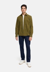Camicia di velluto a coste verde oliva con bottone frontale, taschino sul petto e jeans blu scuro. Il modello indossa sneakers bianche.