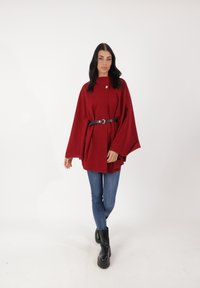 Cappotto stile mantella rosso realizzato in tessuto morbido, con collo alto, bottone frontale e cintura nera. Indossato sopra jeans blu e stivali neri.