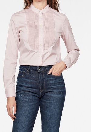 Kvinde iført en lyserød skjorte med knapper og et plisseret frontpanel samt mørkeblå jeans med høj talje, den ene hånd i lommen.