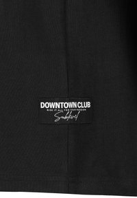 Schwarzes Baumwoll-T-Shirt mit einem genähten Etikett "DOWNTOWN CLUB" in weißem Text und einem charakteristischen Akzent. Sanfte Textur, lässiges Design.