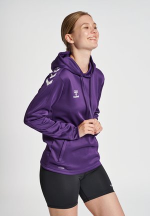 Młoda kobieta uśmiecha się, ubrana w fioletową bluzę z kapturem hummel i czarne sportowe szorty, stojąc na tle jednolitego jasnego tła.