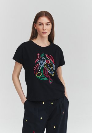 Donna con lunghi capelli castani indossa una maglietta nera con ricamo astratto colorato a foglie e pantaloni scuri con piccoli motivi floreali.