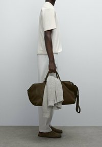 Man i krämfärgad outfit som håller en olivgrön duffelväska med en beige tröja hängande över, stående mot en enkel grå vägg.