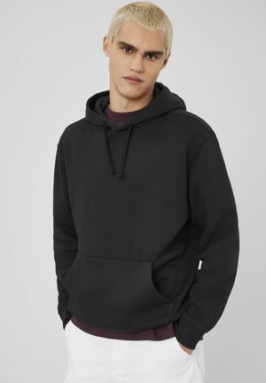 Hoodie noir en mélange de coton, avec une poche avant, capuche ajustable avec cordon de serrage et bords côtes. Porté sur une chemise bordeaux.