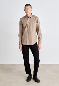 Camisa abotonada de color marrón claro con cuello, ajuste slim, y mangas largas, combinada con pantalones negros y zapatos negro de slip-on.