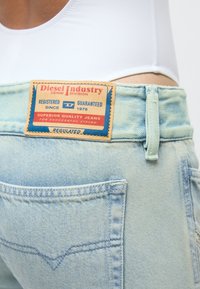 Jean en denim bleu clair avec une étiquette rectangulaire Diesel Industry au-dessus de la poche arrière droite sur une personne portant un haut blanc.