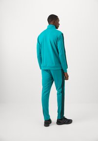 Man standing facing away wearing a turquoise tracksuit with black side stripes and black sneakers on a plain white background.