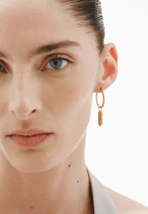 Primer plano de una mujer con ojos azules que lleva un pendiente de aro dorado con un pequeño charm rectangular, sobre un fondo blanco.