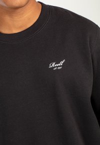 Sudadera negra con cuello redondo acanalado, que presenta el logo bordado en blanco "Reell est. 1997" en el pecho. Textura suave y lisa.