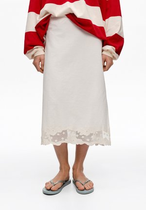 Persona que lleva una falda blanca larga con borde de encaje floral, jersey de rayas rojas y blancas, y sandalias grises tipo chanclas, de pie con los pies juntos.