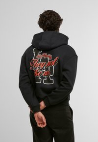 Schwarzer Hoodie mit einem großen Rückendruck, der stilisierte Schriftzüge und ein Buchstabendesign zeigt. Weicher Stoff und lockere Passform, mit Bündchen an den Ärmeln.