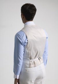 Chaleco de un beige claro con una textura suave, combinado con una camisa de vestir azul. Cuenta con una correa trasera ajustable y detalles de botones.