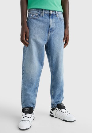 Persoon die lichtblauwe losse jeans, witte en zwarte sneakers en een groen shirt draagt, staand tegen een neutrale achtergrond.