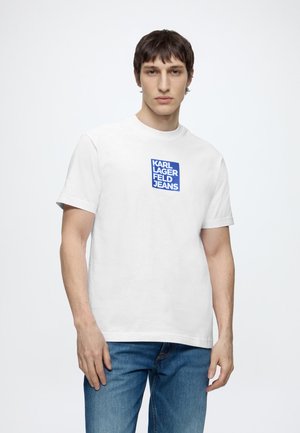 Muž nosiaci biele krátke tričko s modrým logom "KARL LAGERFELD JEANS" a modré džínsy, stojaci pred jednoliatym bielym pozadím.