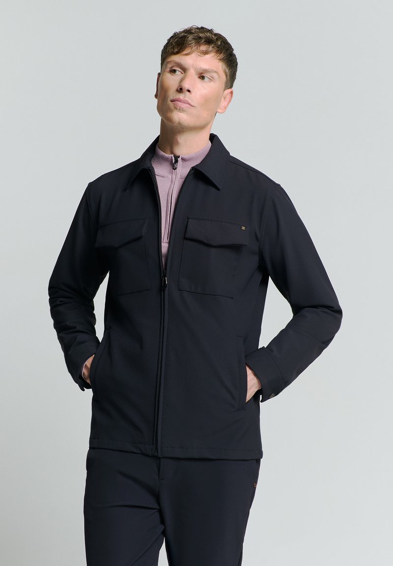 Zwarte ritjacks met twee voorzakken, gemaakt van glad materiaal. Heeft een kraag en een slim fit. Draagt over een lichtpaarse top.