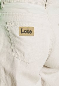 Béžové džínové šortky s texturovaným povrchem. Na zadní kapse se nachází hnědá kožená nášivka s nápisem "Lois", která zdůrazňuje značku.