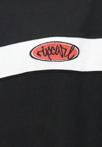 Tecido preto com uma faixa horizontal branca apresenta um emblema oval vermelho com o texto "ripcurl" bordado em preto.