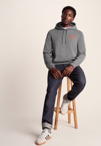 Gray hoodie with a kangaroo pocket and red text detail, paired with dark jeans and white sneakers, seated on a wooden stool.
