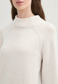 Cremefarbener, gerippter Strickpullover mit hohem Kragen und Raglanärmeln, der über eine weiche Textur und dezente vertikale Streifen verfügt.