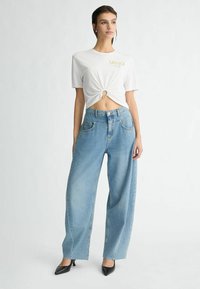T-shirt blanc à manches courtes avec un détail noué et un logo doré, associé à un jean large bleu clair, complété par des escarpins noirs à bouts pointus.