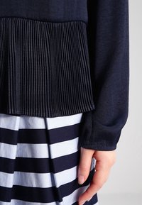 Haut plissé bleu marine avec un corsage texturé, associé à une jupe rayée présentant des bandes horizontales larges alternant bleu et blanc.