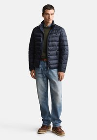 Marinblå vadderad jacka med ståkrage, kombinerad med en grön tröja, blå jeans och bruna stövlar. Slät tyg och quiltad design.