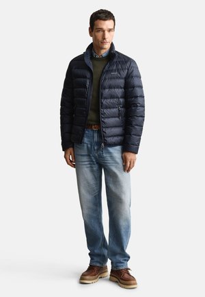 Marineblauwe pufferjas met een opstaande kraag, gecombineerd met een groene trui, blauwe jeans en bruine laarzen. Gladde stof en gewatteerd ontwerp.