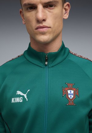 Man wearing teal zip-up sports jacket with Puma logo, "KING" text, and Portugal football crest on front.